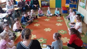 Nauczycielka siedzi na dywanie wraz z dziećmi, które układają działania matematyczne, rodzice obserwują przebieg zajęć siedząc z boku na krzesełkach